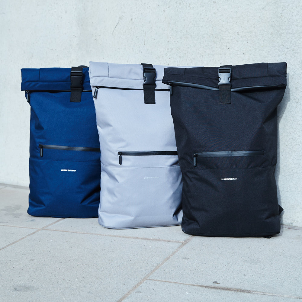 Drei Rolltop Rucksäcke in blau, grau und schwarz stehen vor einer grauen Wand auf dem Boden